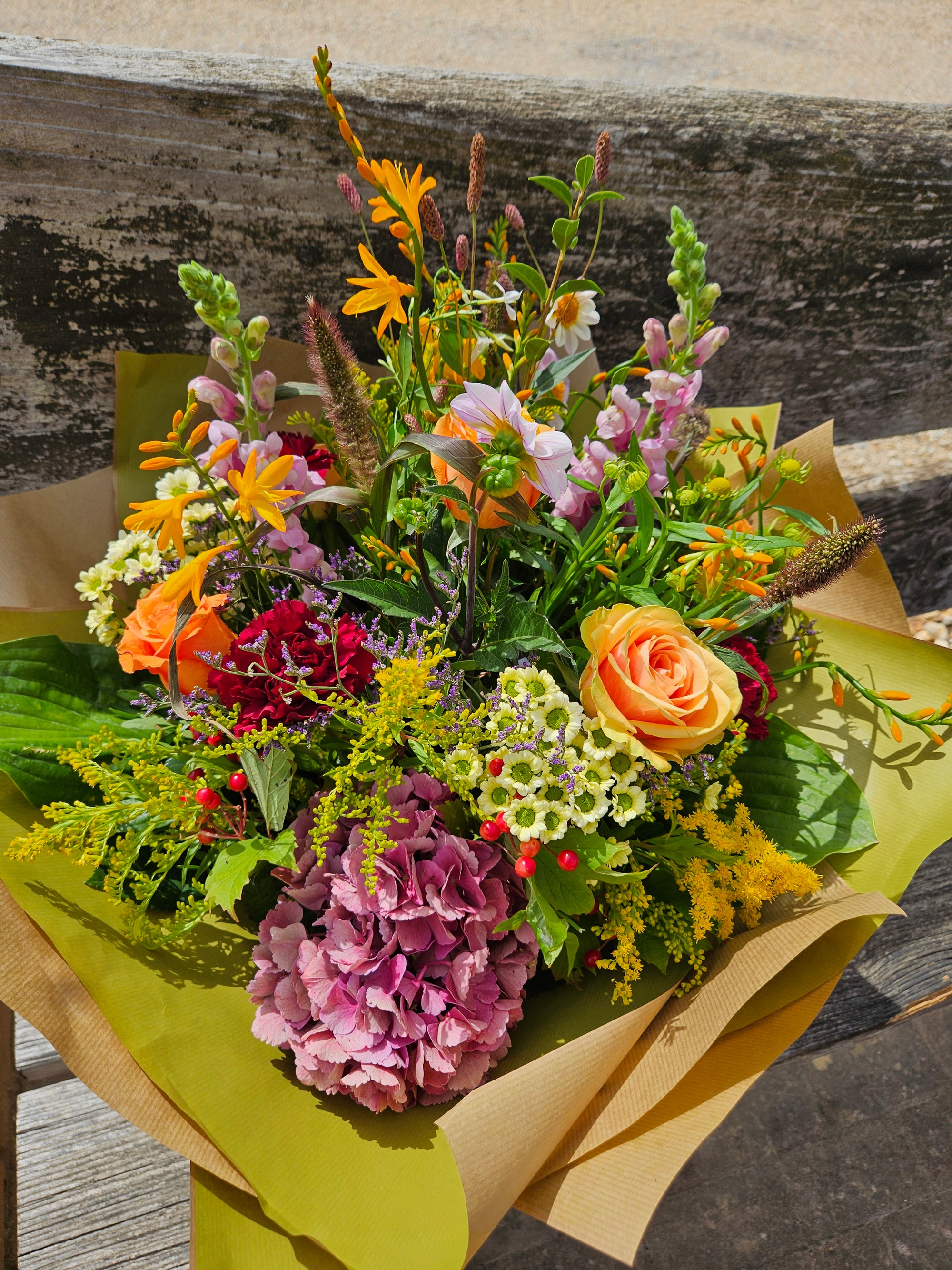 Hand-tied summer bouquet of orange roses, snapdragons and pink hydrangea in kraft wrap on a bench outside our Winchester shop