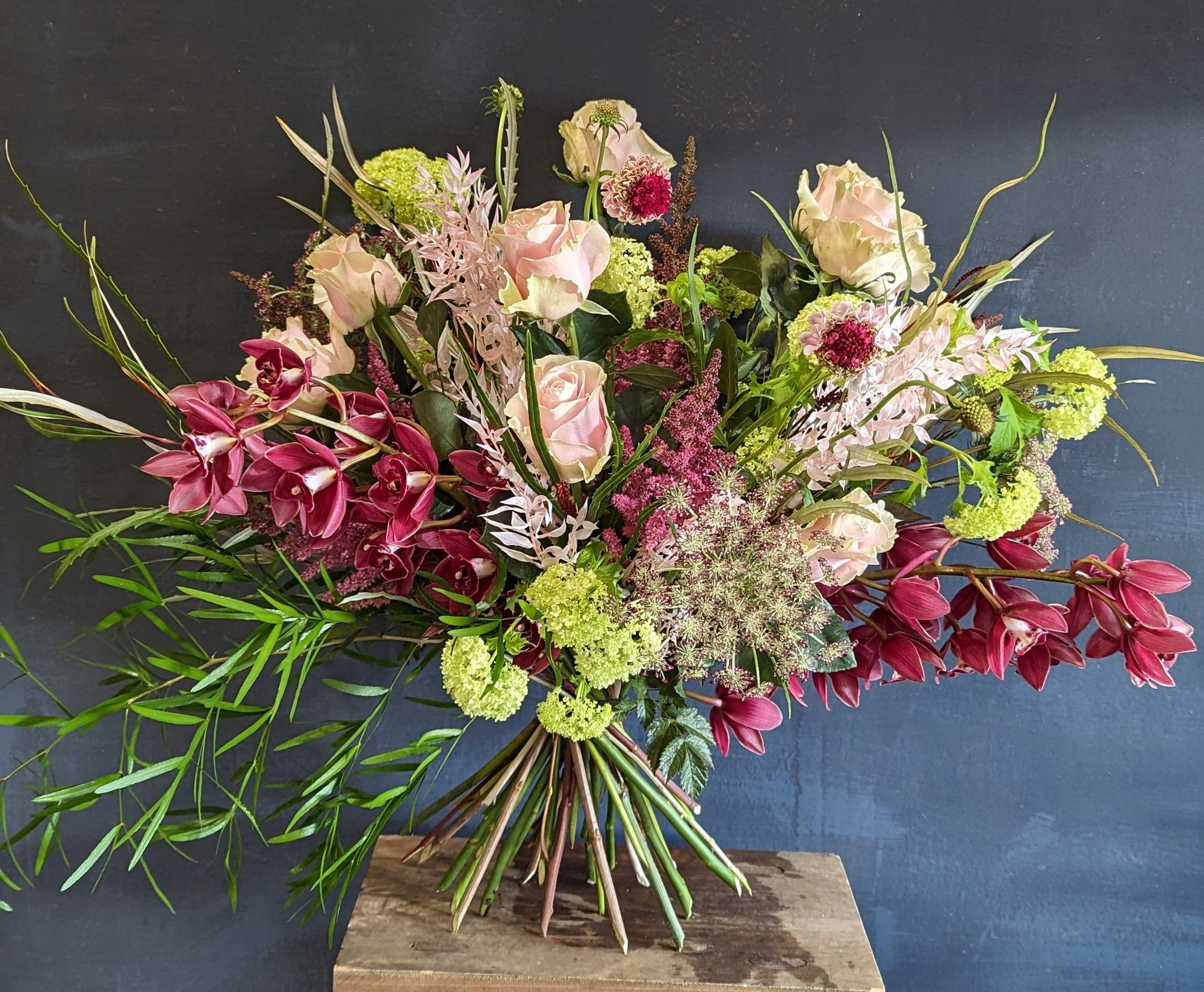 Dramatic hand-tied arrangement of blush roses, burgundy orchids and green viburnum against a dark studio wall